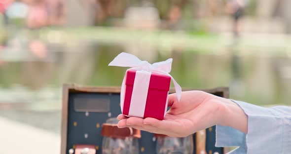 Close Up Women Hand Giving Red Box with White Silk Ribbon in Green Urban Park