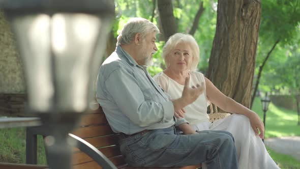 Side View of Relaxed Retirees Chatting and Laughing in Sunny Park. Senior Caucasian Husband and Wife