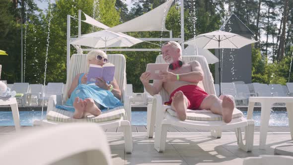 Adorable Positive Couple Lying on Sunbeds Near the Pool. The Woman Reading the Book