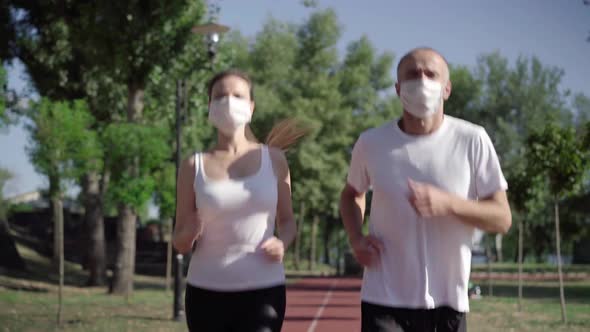 Serious Caucasian Man and Woman in Face Masks Jogging During Coronavirus Viral Pandemic Outdoors