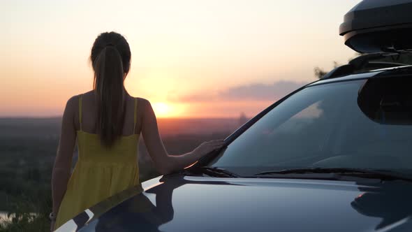 Happy young girl in summer dress standing near her car enjoying sunset view. Travel and vacations 