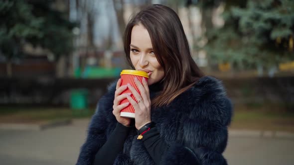Long-haired girl with plastic cup outdoors. Attractive young woman in fur coat enjoys drinking coffe