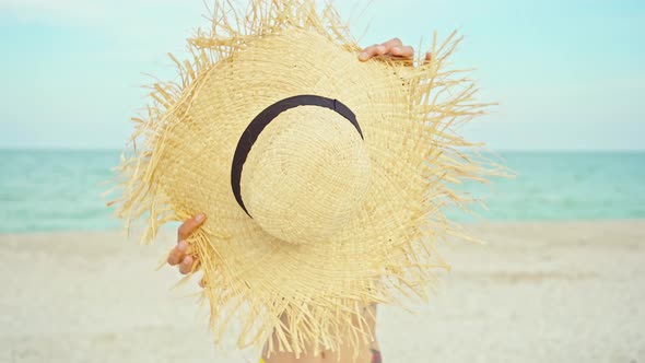 Woman Holding Summer Straw Hat in Front to Camera on Windy Ocean Beach