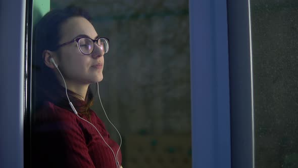 A Young Woman Is Sitting at Home in Quarantine with Headphones in Her Ears. A Girl Sits on a Window