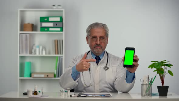 Aged Male Doctor Holds Smartphone with Green Screen Chroma Key Points at It with Finger