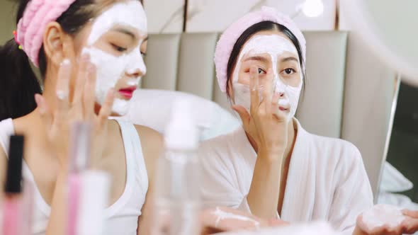 Beautiful young woman in white bathrobe applying a revitalizing  mask her friend's face.