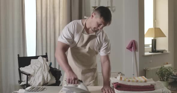 Portrait of Diligent Caucasian Young Man Thoroughly Ironing Bed Linen Indoors, Handsome Brunette Guy