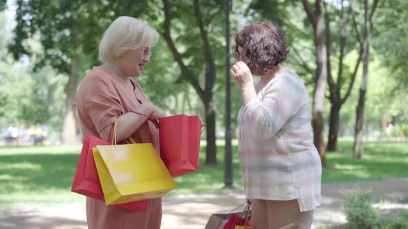 Two Cheerful Female Retirees Bragging Purchases in Sunny Park. Side View Portrait of Happy Smiling