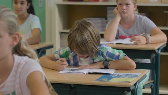 Young boy drawing in classroom while the rest of the class is paying attention - wide