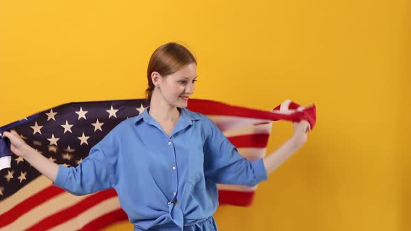 Young Beautiful Woman in Bright Clothes on a Pastel Yellow Background Holds and Develops a USA Flag