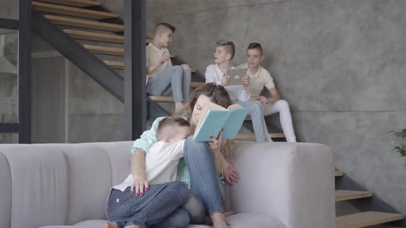 Pretty Young Mother Reading Book To Her Son While the Rest of Her Teen Kids Playing with Each Other
