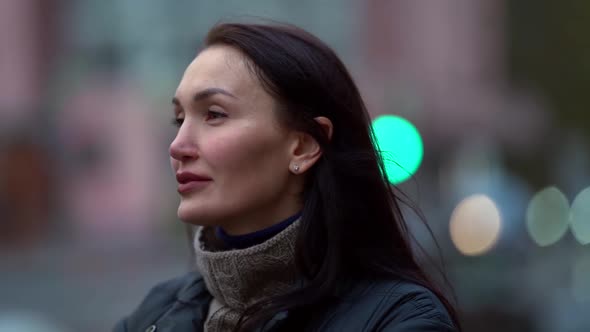 a Portrait of a Brunette with Long Hair in a Knitted Sweater and a Dark Coat on a Cloudy Day Against