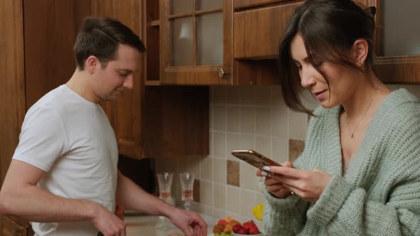 Young Man Prepares Food in the Kitchen While His Wife Is Surf the Internet