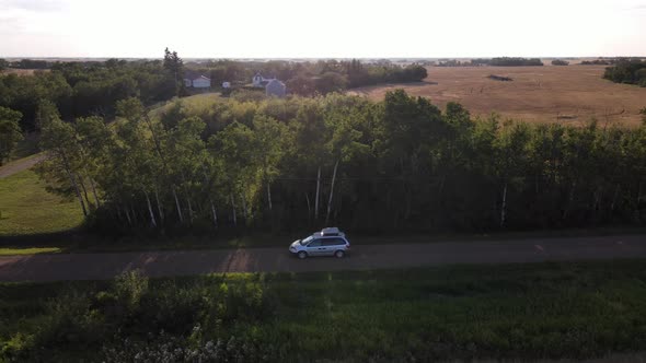 4k aerial shot of silver minivan driving slowly along dirt road next to trees during sunset in rural
