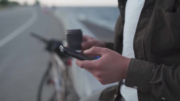 Unrecognizable Male Cyclist Use Smartphone and Drinks Takeaway Coffee While Standing Near Bike on