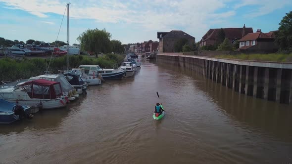 Man kayaking on meandering river next to boats and houses, drone closely follows and pans up to reve