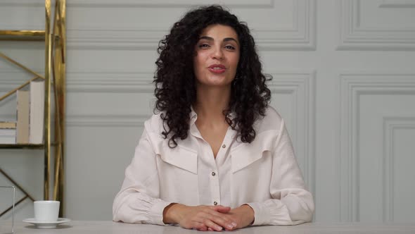 Young Indian Woman with Curly Hair Talking to a Camera with a Friend Online Sitting at a Desk in the