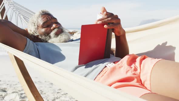 Happy senior african american man reading and lying in hammock on sunny beach