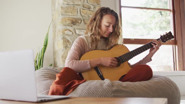 Happy caucasian woman playing acoustic guitar, sitting on beanbag in sunny cottage living room