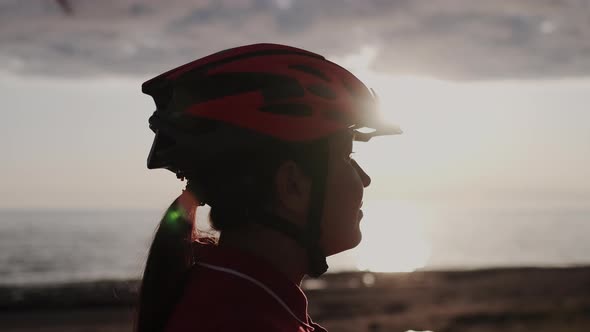 Young Asian Female Cyclist Checking Red Protective Helmet at Sunset Time with Sun Beams in Camera