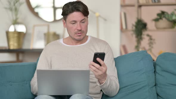 Middle Aged Man with Smartphone Working on Laptop, at Home