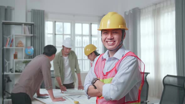 Asian Man Engineer With Helmet Crossing Arms And Smiling To Camera While His Colleagues Working