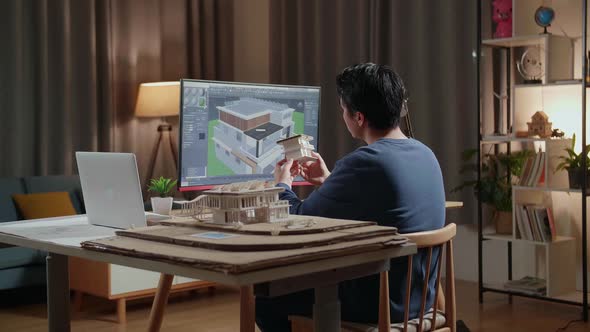 Male Engineer Holding And Looking At House Model With Solar Panel While Designing House On A Desktop