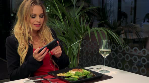 A Young Woman Sits at a Table in a Restaurant and Takes a Picture of a Salad with Her Smartphone