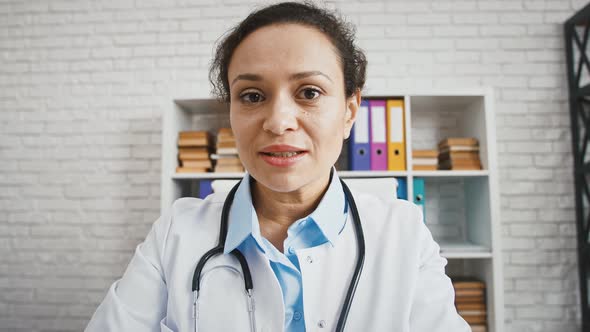 Professional Female Doctor in White Uniform with Stethoscope Sitting at Office Talking to Camera
