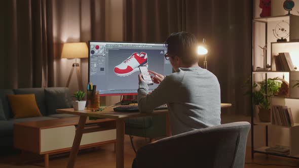 Asian Male Footwear Designer Looking At Photos On Smartphone While Designing Shoe On A Desktop