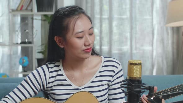 Close Up Of Asian Woman With Microphone Having Video Call On Laptop While Playing A Guitar At Home
