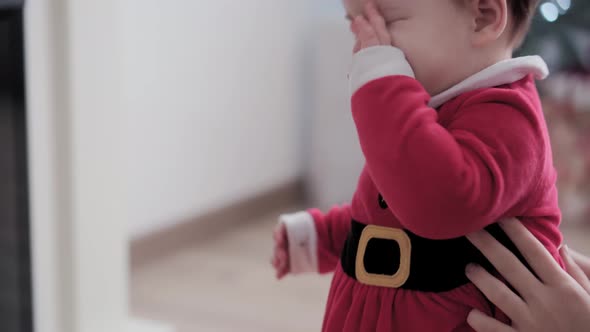 Authentic Cute Chubby Infant Baby Girl in Santa Hat and Red Dress Near Fireplace with Young Woman