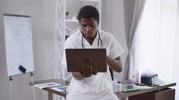 Focused Intelligent African American Male Doctor with Laptop Surfing Internet Sitting on Table in