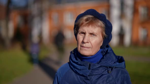 Portrait of an Elderly Woman in a Beret and Blue Clothes Against the Background of Blurred Houses on