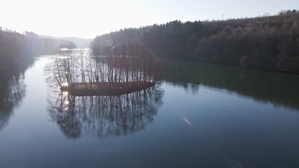 Aerial fly by shot of a tiny island covered by leafless trees at sunset.