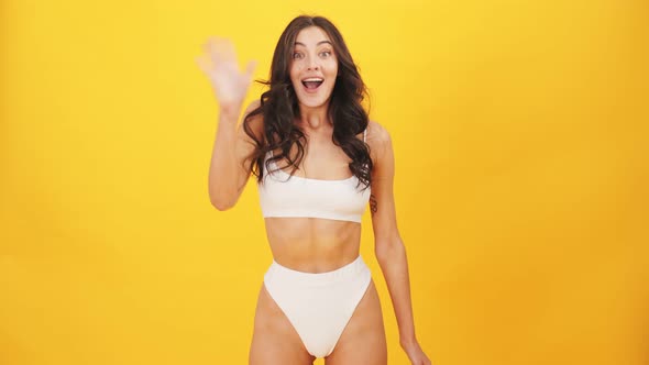 A smiling brunette woman wearing swimsuit doing hello gesture