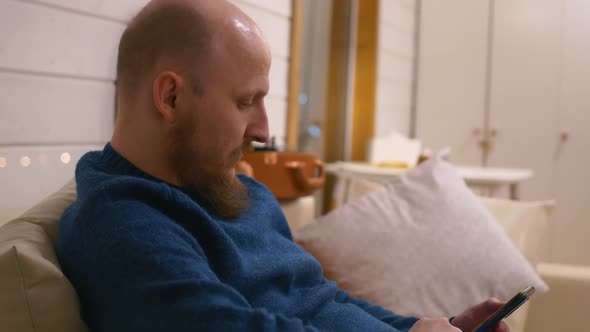 A Bald Man with a Beard Sits on the Couch at Home in the Evening and Browses Social Networks in a