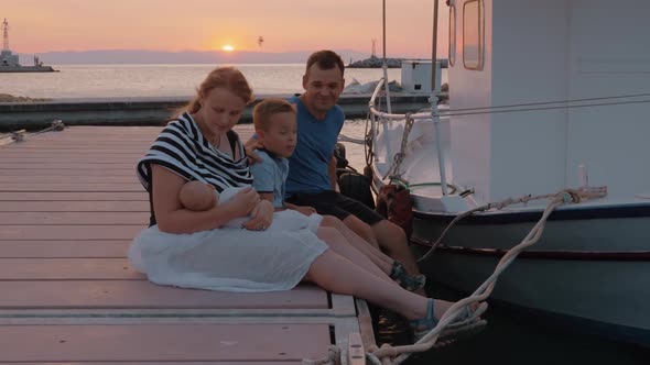 Parents with Elder Son and Baby Sitting on the Pier at Sunset