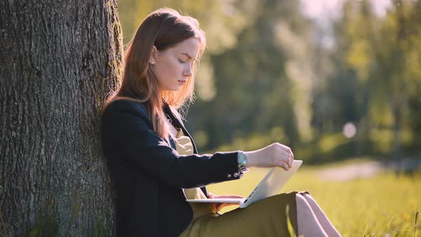 A Girl with a Laptop Resting in the Park By a Tree