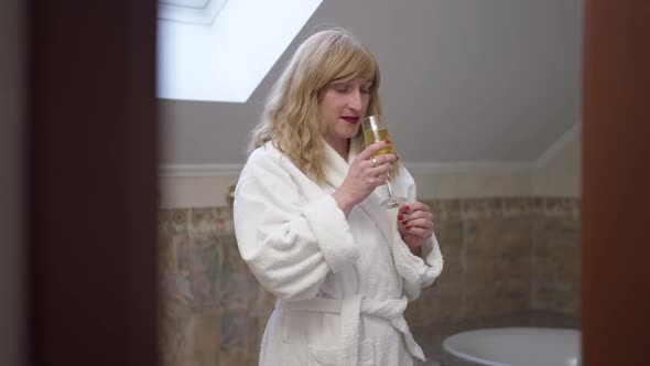 Relaxed Carefree Transgender Woman in Bathrobe Enjoying Taste of White Wine Standing in Bathroom