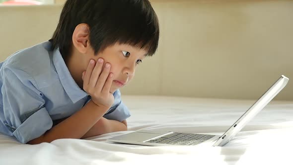 Cute Asian Boy Lying In Bed And Using Laptop On White Bed