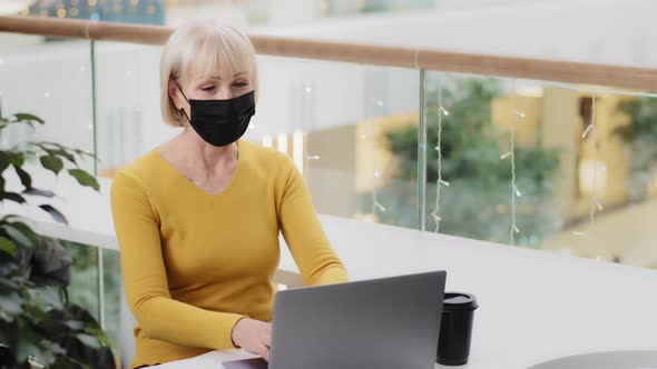 Elderly Woman in Protective Mask Sits at Desk Typing on Laptop Playful Excited Female Player Plays