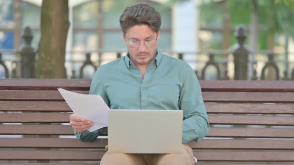 Successful Middle Aged Man Reading Documents while using Laptop