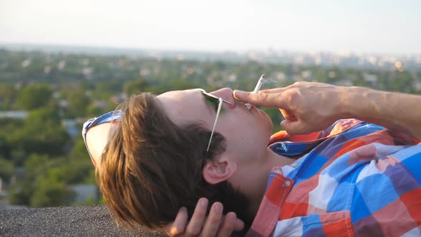 Close Up of Handsome Man in Sunglasses Lying on Roof and Smoking Cigarette at Cityscape Background