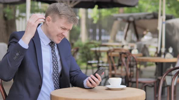 Businessman Upset by Loss on Smartphone, Sitting in Outdoor Cafe