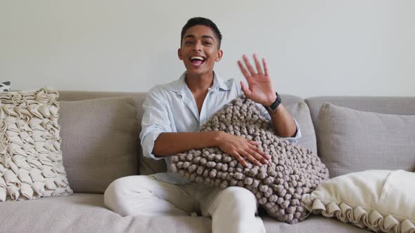 Happy mixed race man sitting on sofa holding cushion laughing and waving