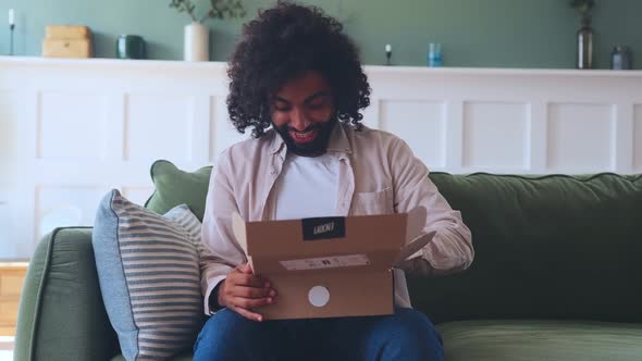 Young Arabian Man with Smile Unpack Cardboard Box Sits on Sofa in Living Room