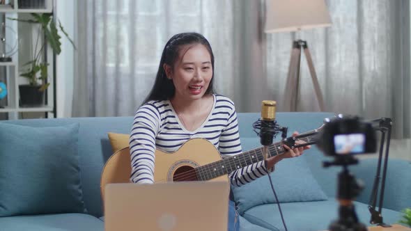 Asian Woman Reading Comments On Laptop And Speaking To Camera While Live Stream Playing A Guitar