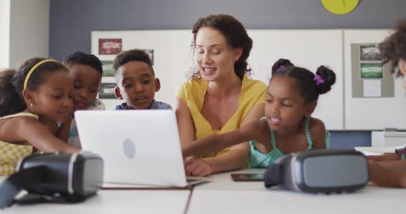 Video of happy caucasian female teacher and african american pupils using laptop and vr headsets alt