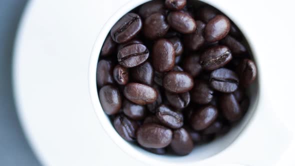 Roasted Coffee Beans Being Poured Into White China Cup On Saucer Overhead View